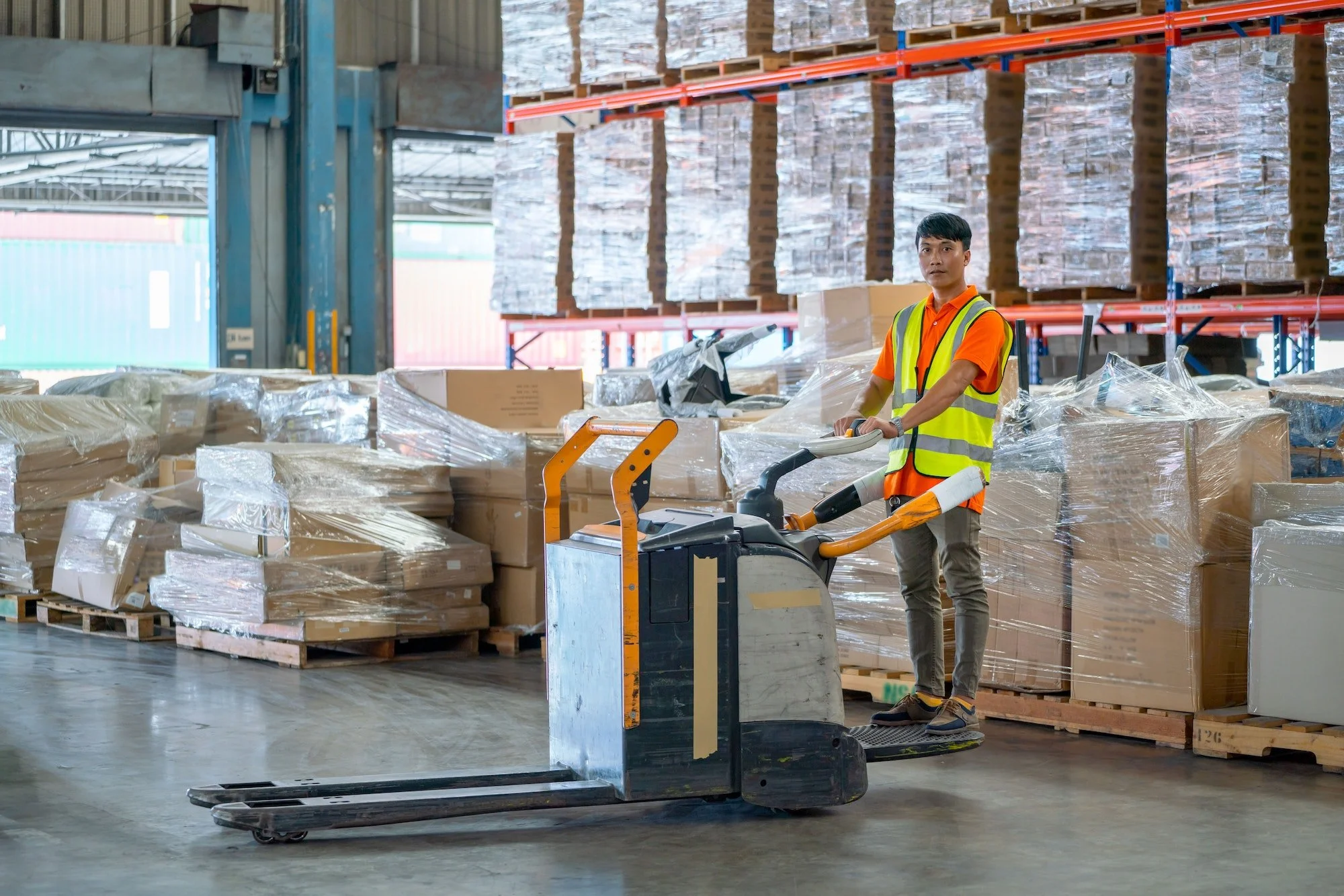 Asian warehouse worker stand and drive folklift in front of stack of product carton in workplace