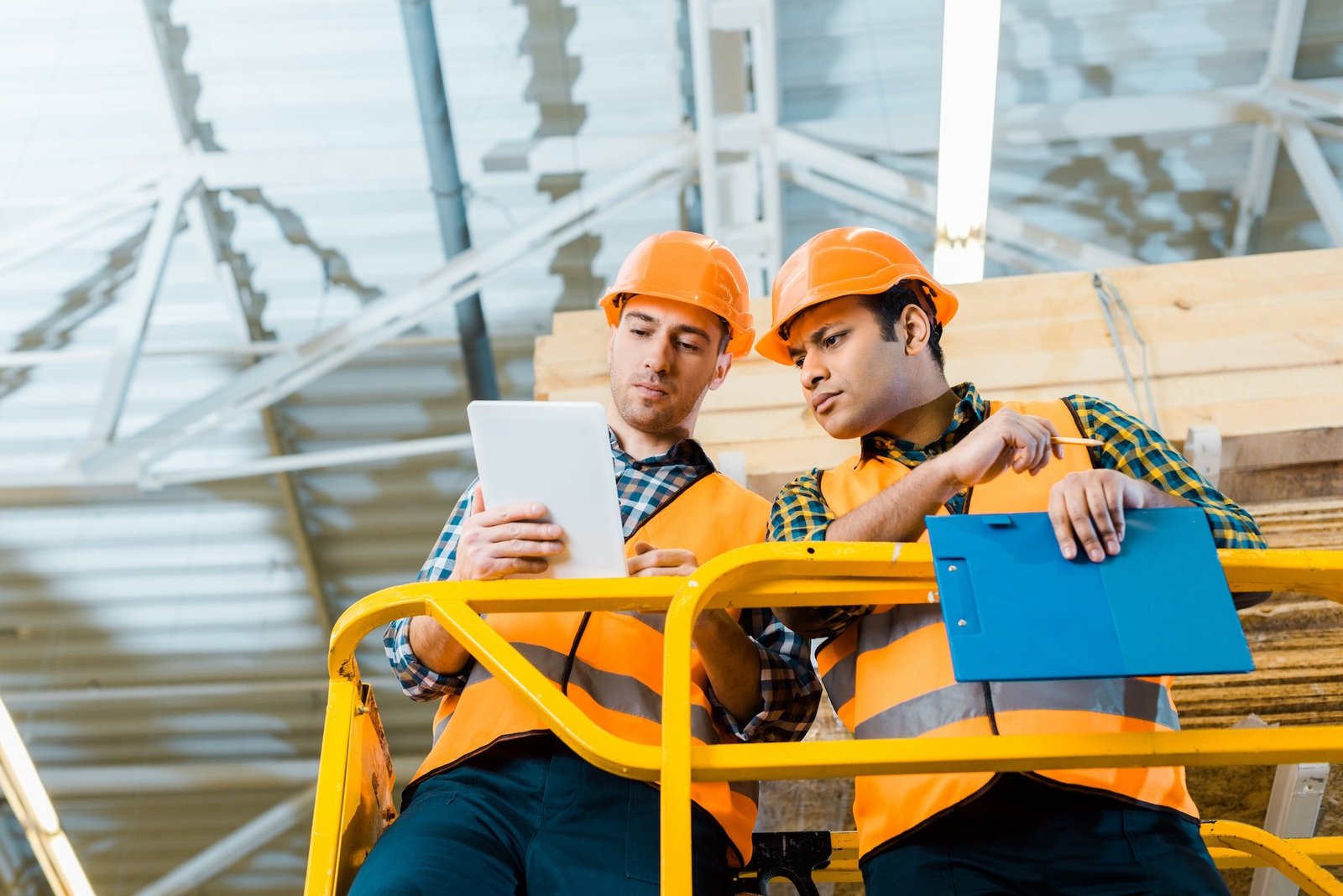 thoughtful multicultural workers using digital tablet while standing on scissor lift in warehouse